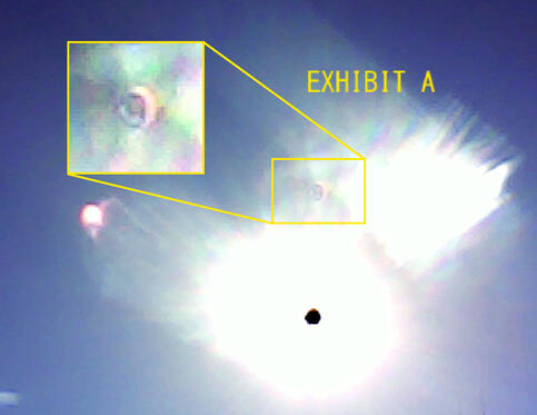 a low-resolution photograph of the sky, containing a large sun glare and what appears to be a wide-open eyeball with a blue iris. a yellow-outlined box contains an enlargement of the eyeball-like glare, and yellow text next to it reads "exhibit A." this im